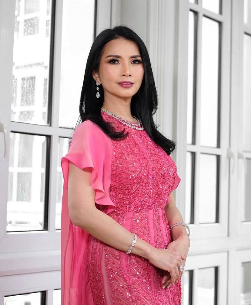 Seorang wanita berambut hitam panjang mengenakan gaun merah muda berhias payet dan perhiasan elegan berdiri di depan jendela besar.