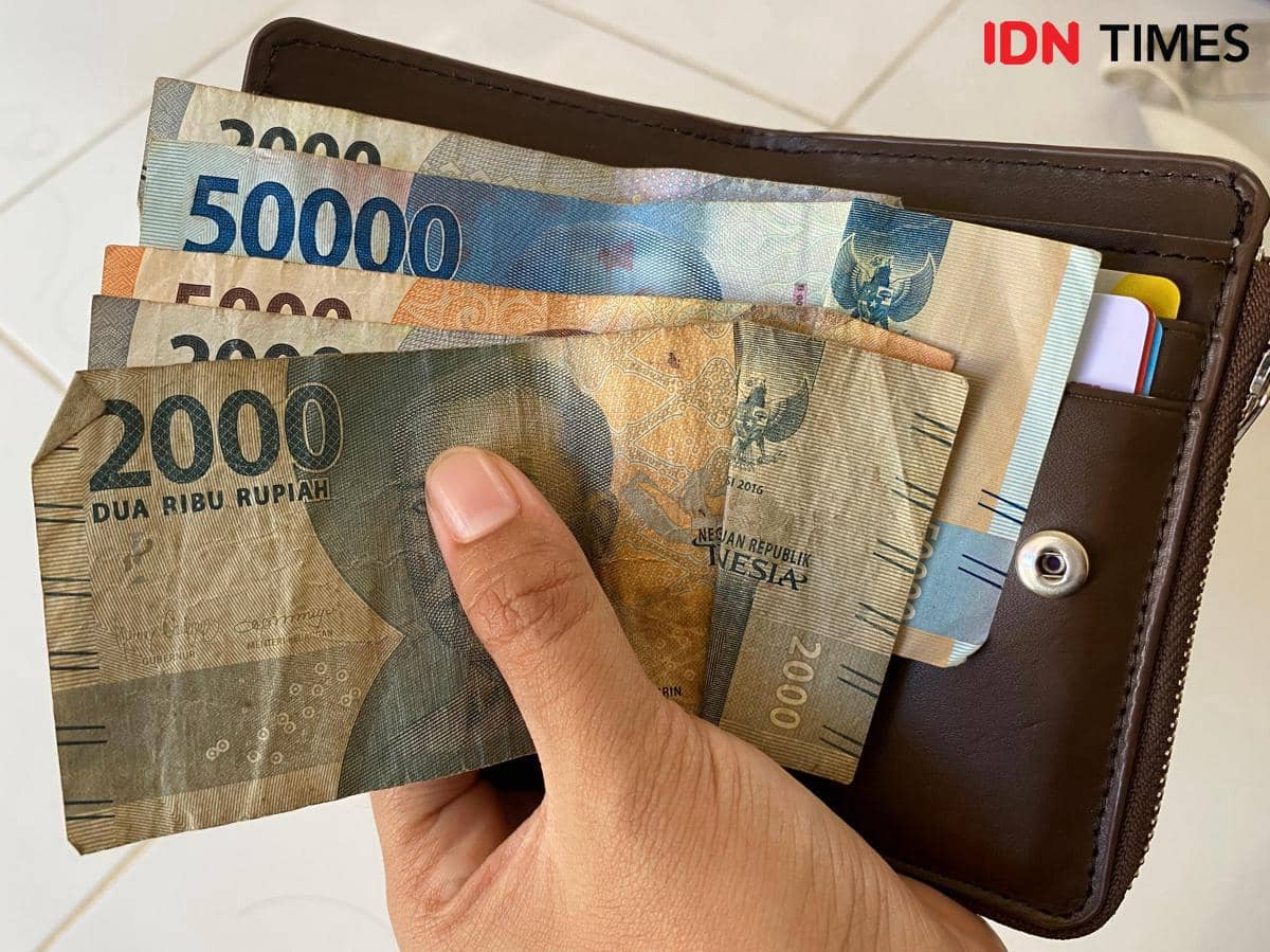 Tangan memegang dompet cokelat berisi uang kertas rupiah pecahan 2.000, 5.000, dan 50.000 di atas permukaan terang.