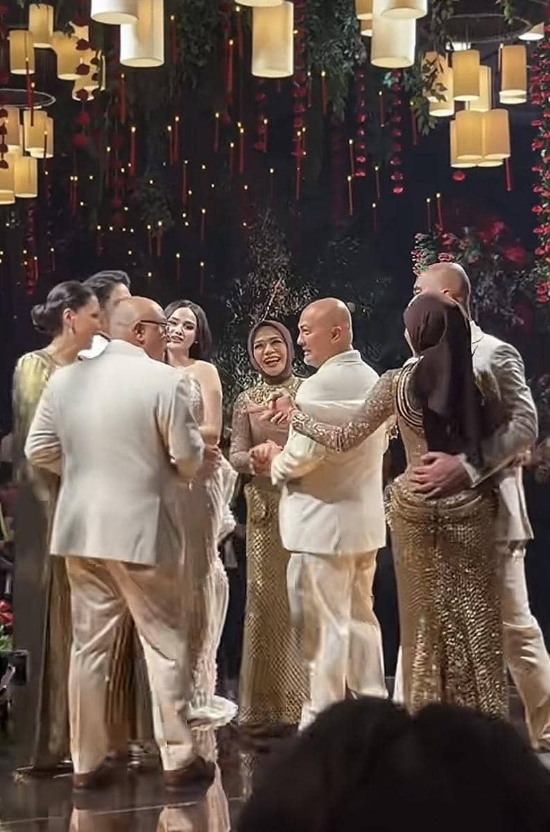 Beberapa pasangan mengenakan busana formal berwarna emas dan putih sedang berdansa di acara pesta dengan dekorasi lampu gantung dan bunga.