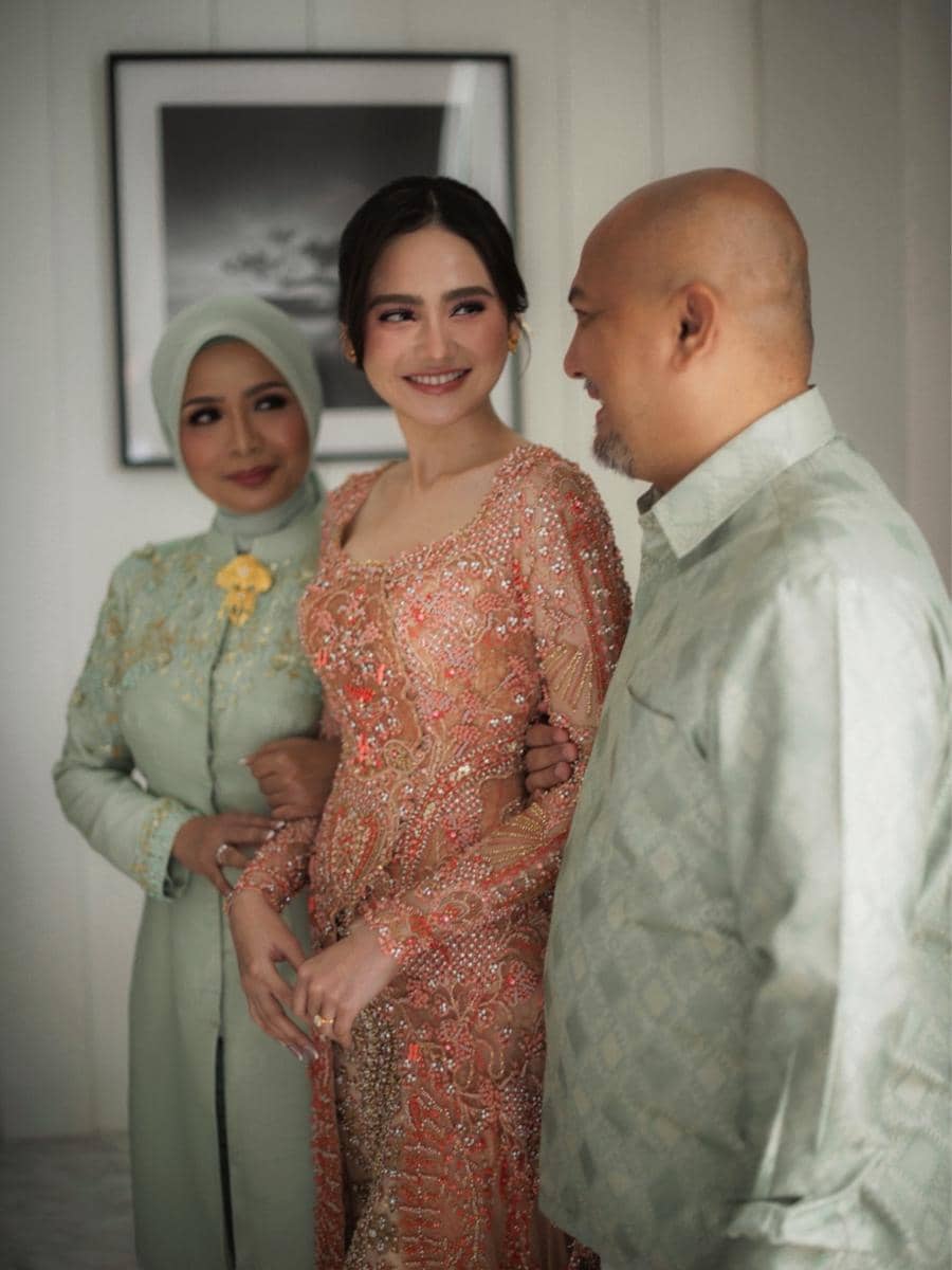 Seorang perempuan mengenakan kebaya berwarna oranye berhias payet berdiri di antara pria dan wanita berpakaian hijau pastel sambil tersenyum hangat.