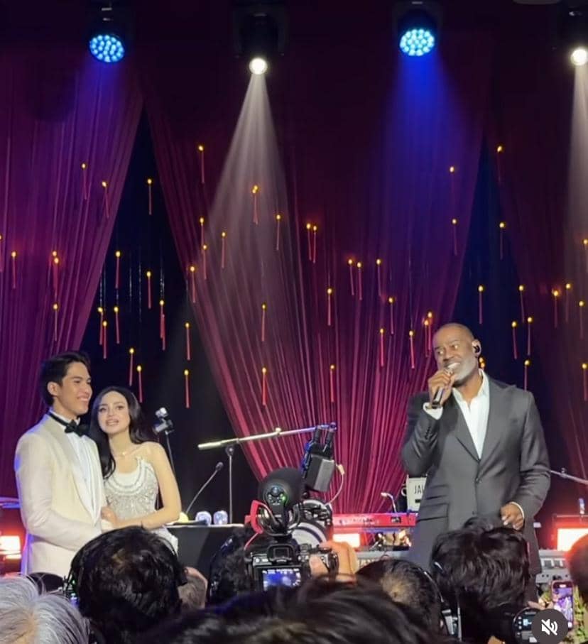 Seorang penyanyi tampil di atas panggung dengan mikrofon di tangan, sementara sepasang pengantin berdiri tersenyum di sampingnya di resepsi pernikahan.