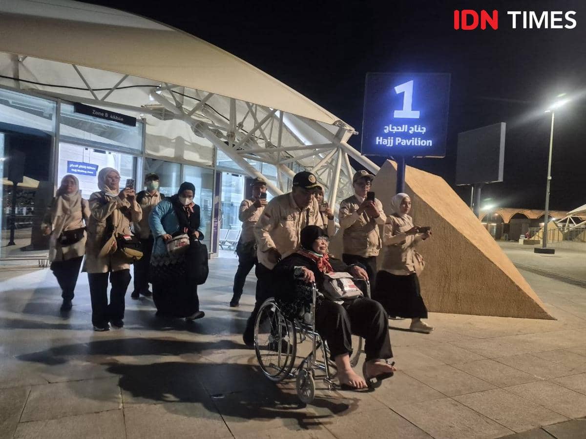 Petugas PPIH Arab Saudi mendorong jemaah haji lansia di kursi roda menuju bus khusus di area Hajj Pavilion pada malam hari.