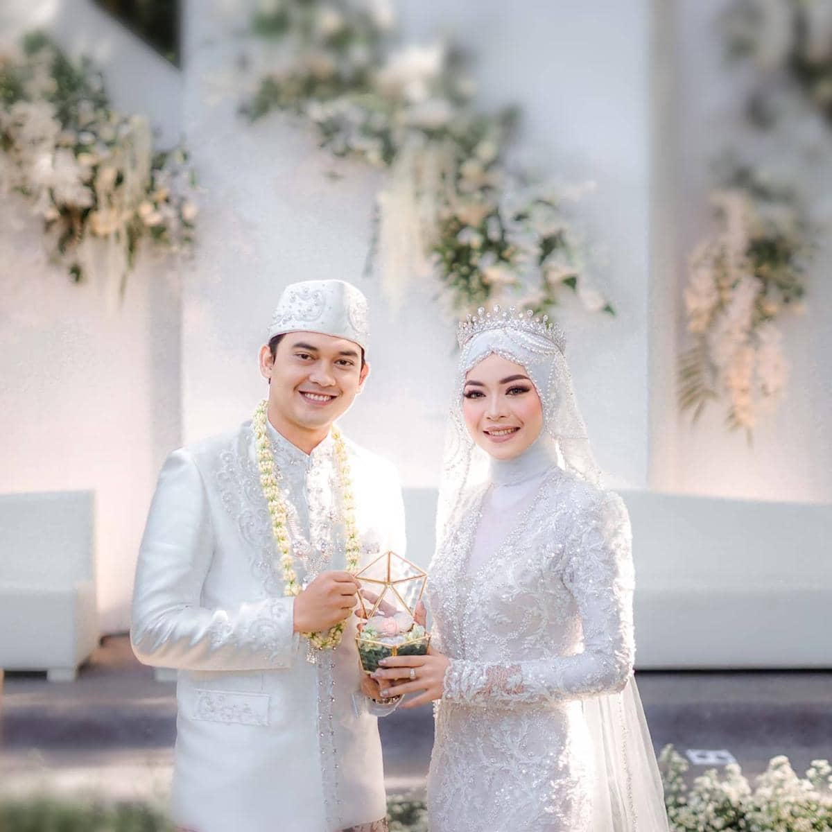 Sepasang pengantin mengenakan busana adat berwarna putih berhias detail bordir, tersenyum sambil memegang wadah dekoratif di pelaminan.