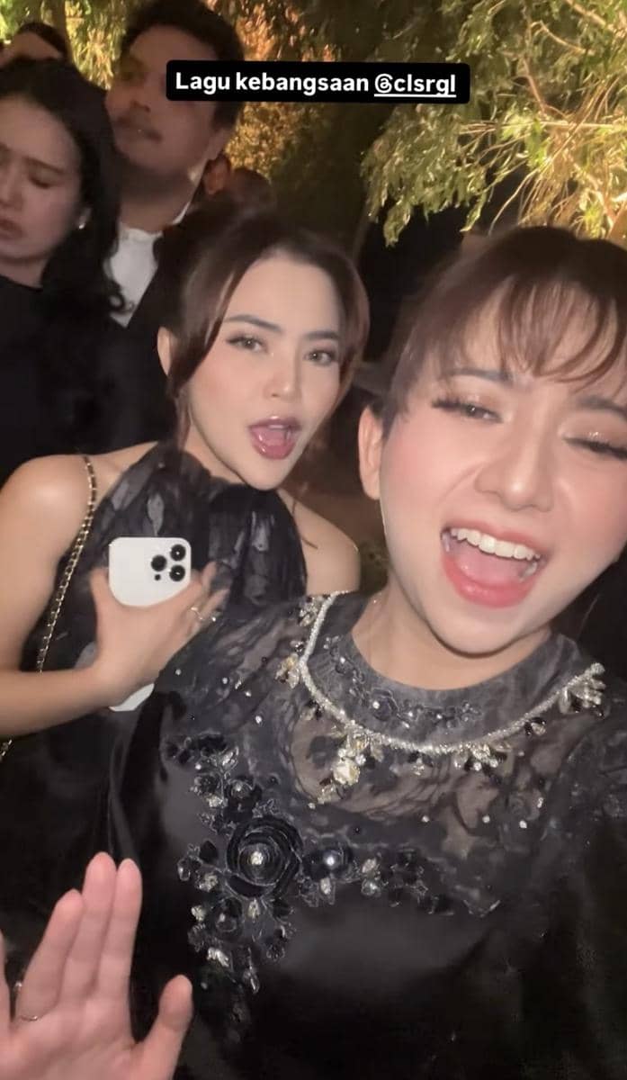 Dua wanita mengenakan gaun hitam berpose sambil tersenyum dan bernyanyi di acara pernikahan pada malam hari.