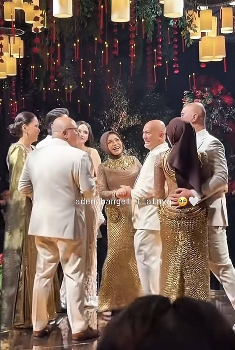 Beberapa pasangan mengenakan busana formal berwarna emas dan putih sedang berdansa di acara dengan dekorasi lampu gantung dan nuansa hangat.