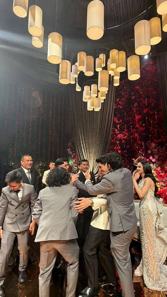 Para groomsmen mengenakan setelan abu-abu sedang bercanda di acara pernikahan dengan dekorasi lampu gantung dan bunga merah di latar belakang.