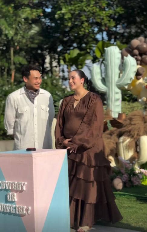 Seorang wanita hamil mengenakan gaun cokelat berdiri di samping pria berbusana putih dalam acara gender reveal bertema alam terbuka.