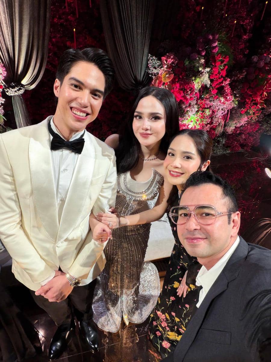 Empat orang berpakaian formal berpose bersama di resepsi pernikahan dengan latar dekorasi bunga merah dan tirai elegan.