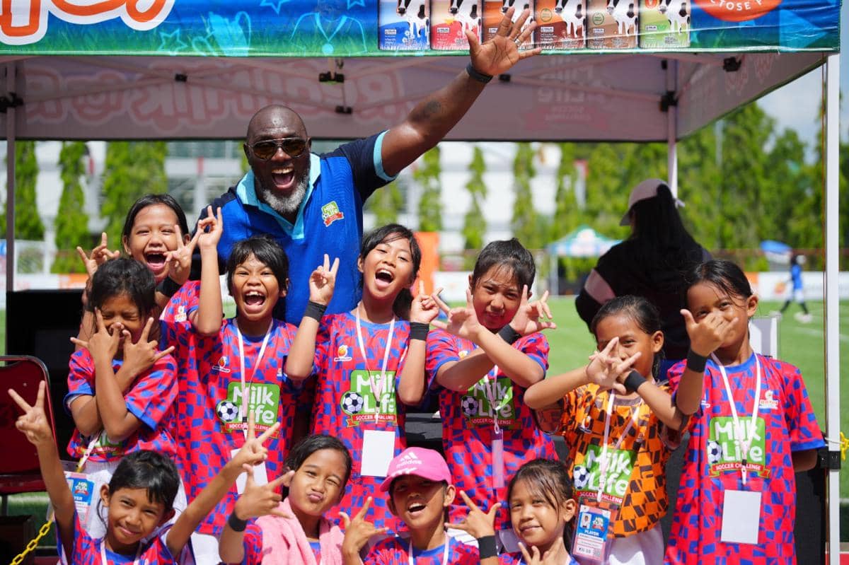 Seorang pelatih berpose gembira bersama sekelompok anak-anak peserta MLSC Samarinda Seri 2 2025-2026 mengenakan seragam sepak bola berwarna cerah.