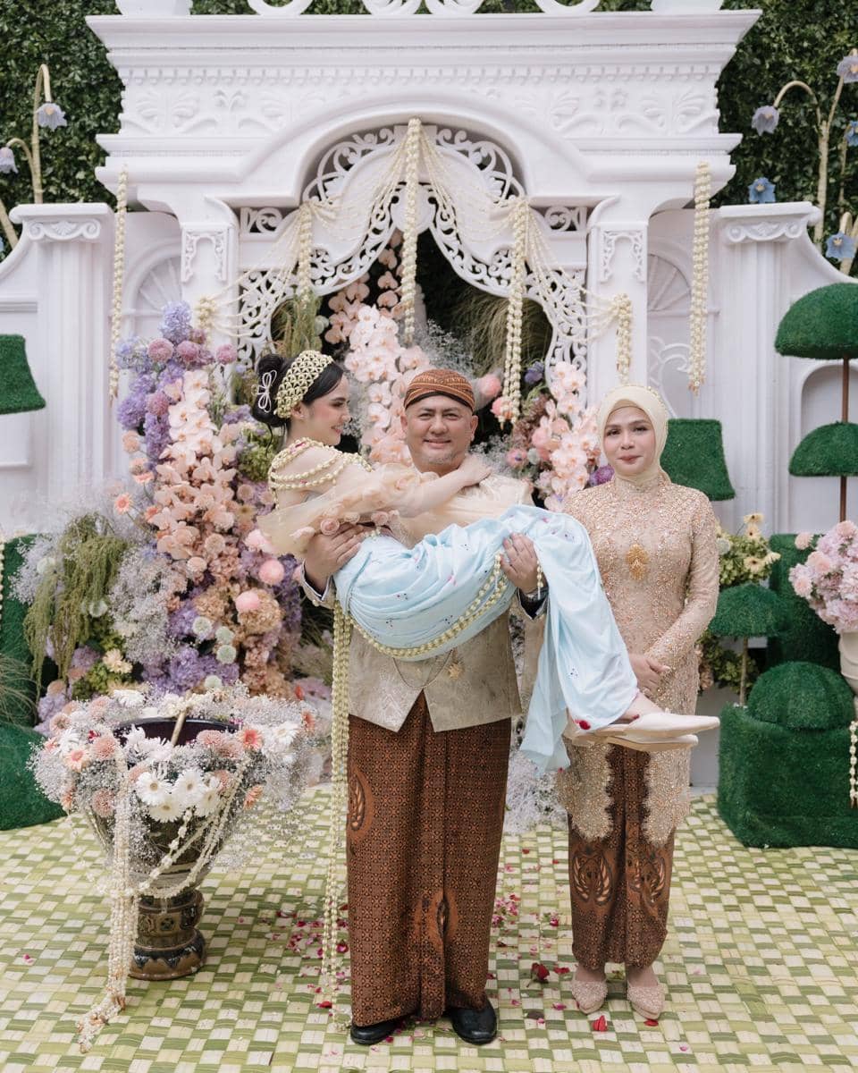 Seorang pria mengenakan pakaian adat Jawa menggendong seorang wanita dalam balutan busana tradisional di acara pernikahan dengan latar dekorasi bunga berwarna pastel.