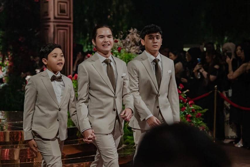 Tiga pria mengenakan setelan jas krem dengan dasi cokelat berjalan bersama di acara resepsi pernikahan yang mewah.