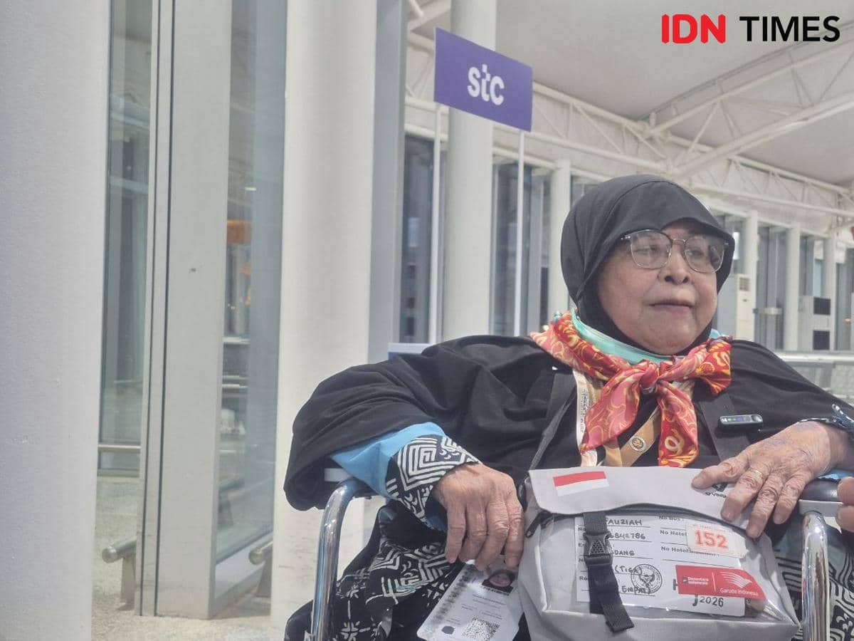 Seorang jemaah haji lanjut usia mengenakan pakaian ihram dan duduk di kursi roda di area bandara dengan tas bertanda identitas haji.