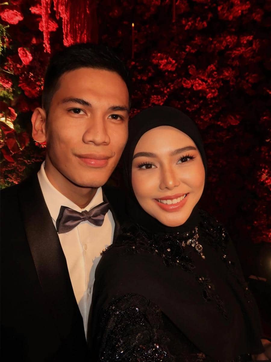 Sepasang tamu berpakaian formal berpose bersama di acara resepsi dengan latar bunga merah yang mewah dan pencahayaan hangat.