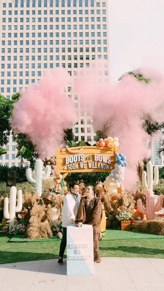 Sepasang calon orang tua berdiri di depan dekorasi pesta gender reveal bertema koboi dengan asap berwarna merah muda di latar belakang.