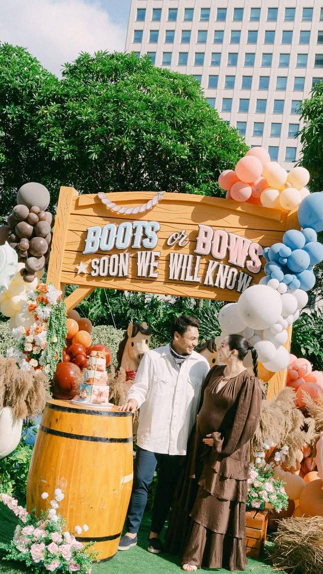 Pasangan berdiri tersenyum di acara gender reveal bertema western dengan dekorasi balon warna-warni dan papan bertuliskan 'Boots or Bows'.