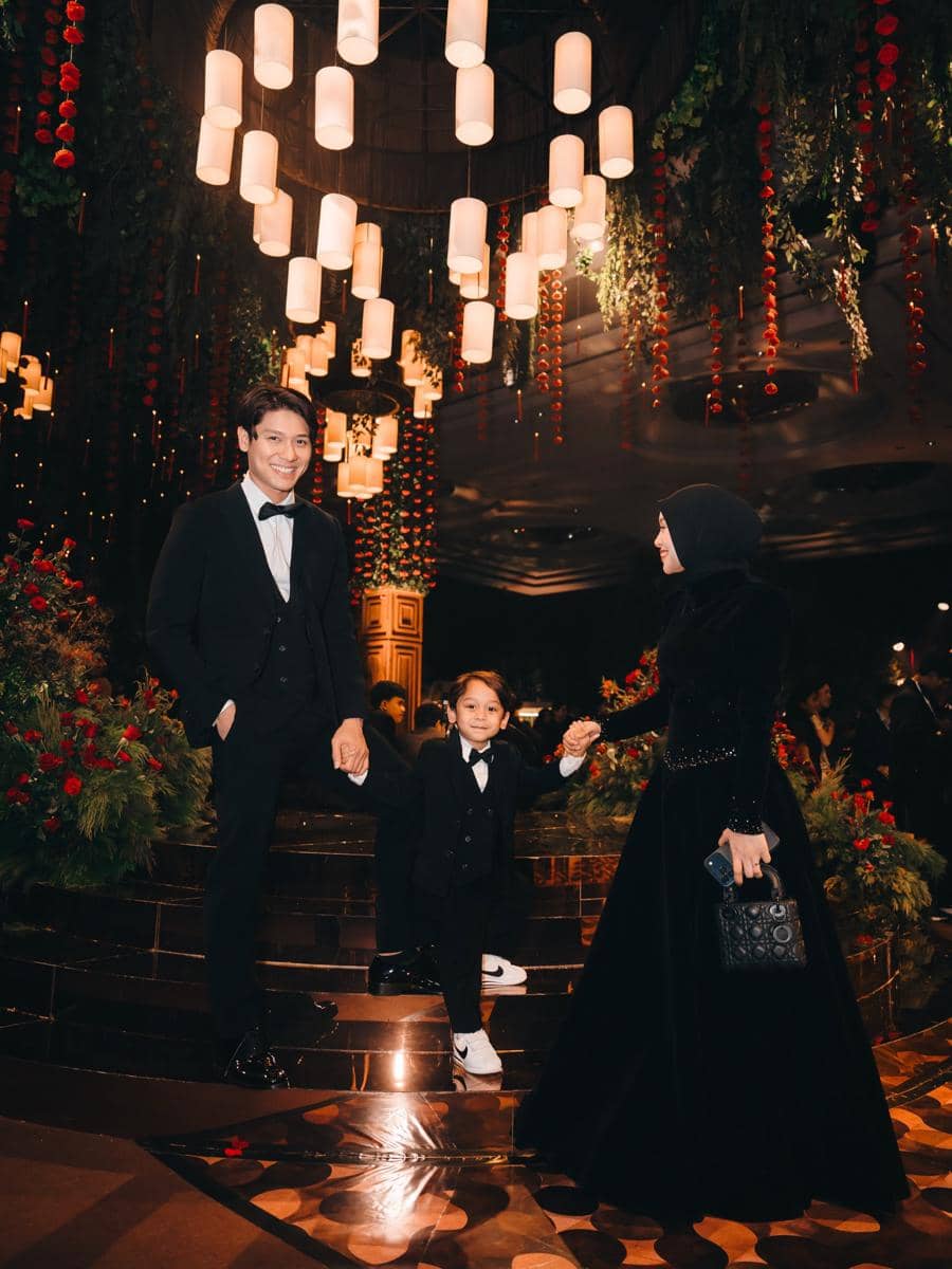 Sebuah keluarga berpakaian formal hitam berpose di tangga dengan dekorasi lampu gantung dan bunga merah di resepsi pernikahan.