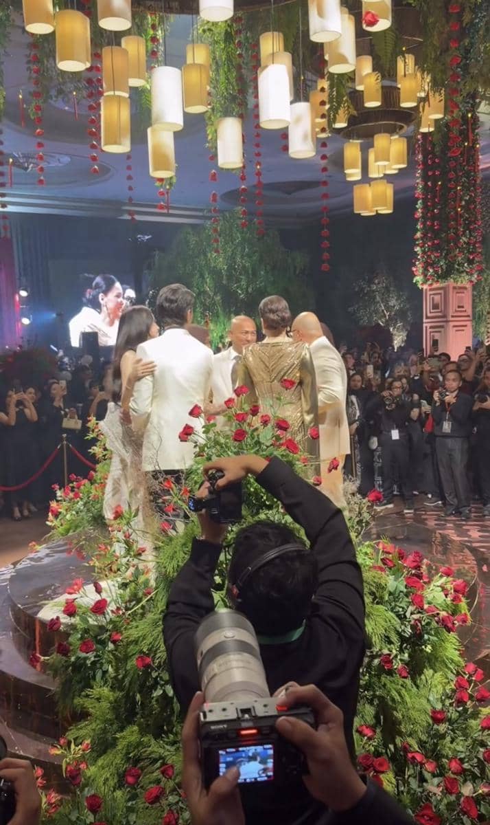 Suasana pesta pernikahan mewah dengan pasangan dan orang tua berdansa di panggung dikelilingi dekorasi bunga mawar merah dan lampu gantung elegan.