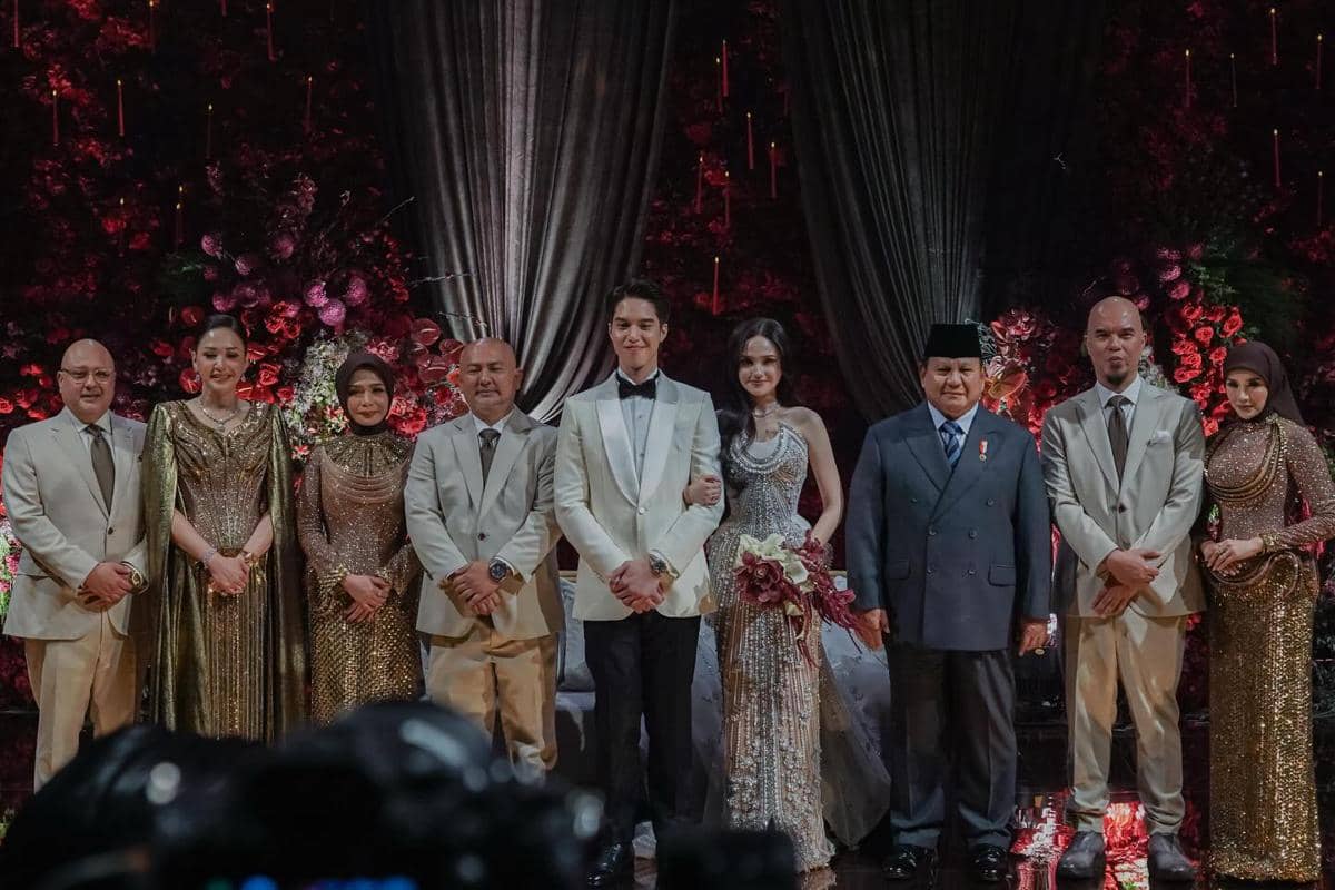 Sejumlah tamu dan keluarga berpose bersama pengantin di acara pernikahan dengan latar dekorasi bunga merah dan tirai elegan.