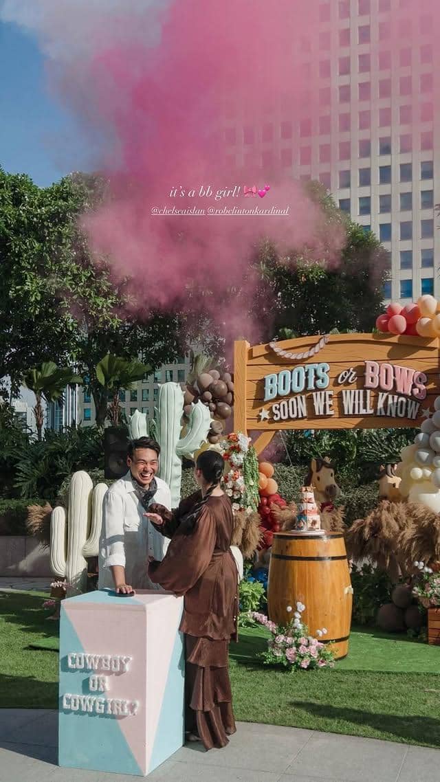 Pasangan merayakan pesta gender reveal bertema koboi dengan asap berwarna merah muda di area luar ruangan yang dihiasi balon dan dekorasi kaktus.