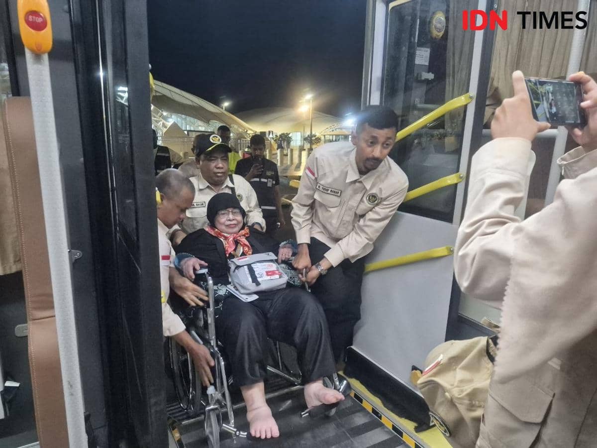 Petugas berseragam membantu seorang jamaah perempuan lanjut usia di kursi roda naik ke bus khusus di area bandara pada malam hari.