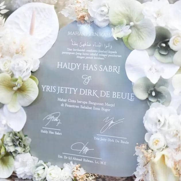 Mahar pernikahan berdesain elegan bertuliskan nama pasangan Haldy Has Sabri dan Yris Jetty Dirk De Beule, dihiasi bunga putih dan hijau lembut.