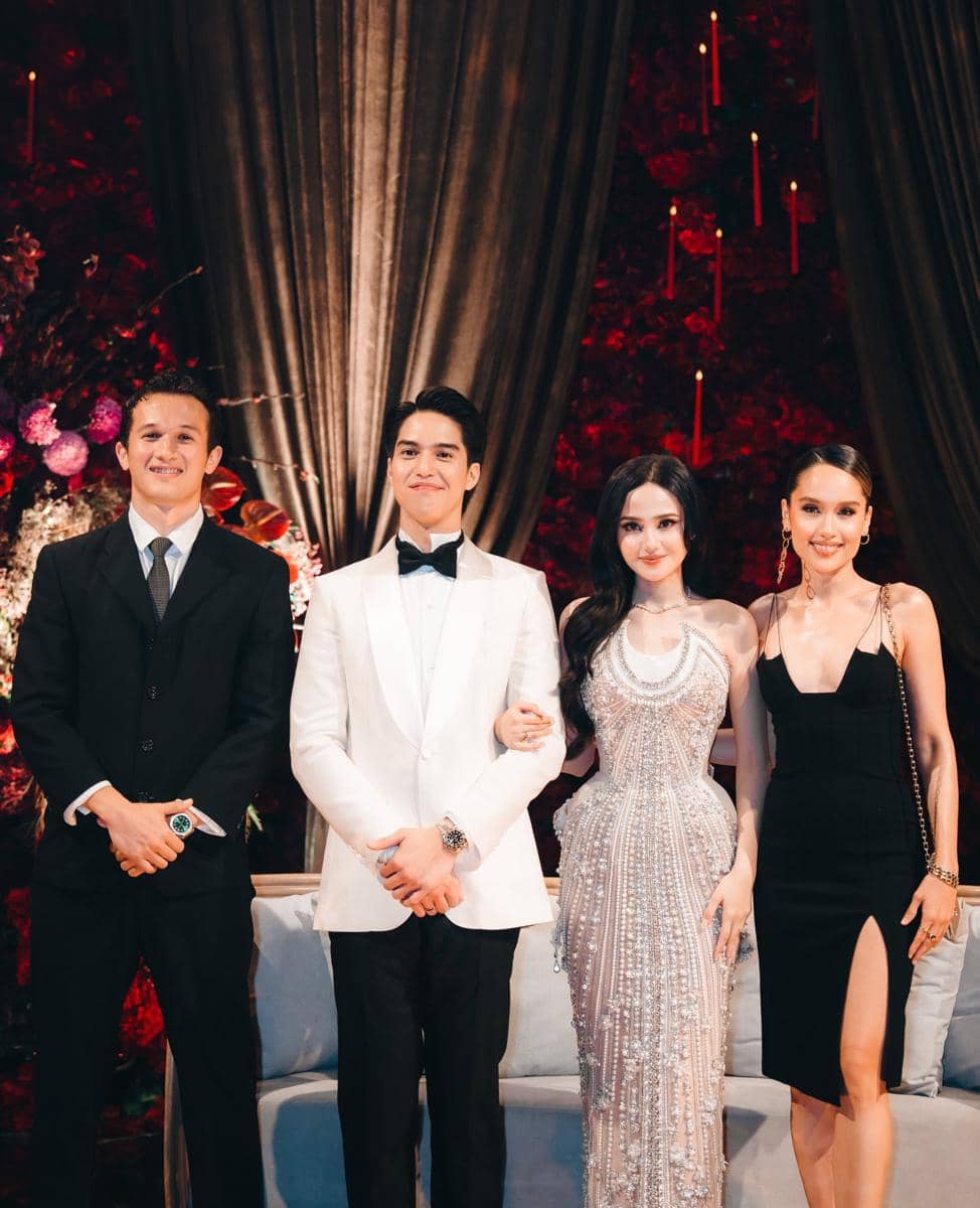 Empat orang berpakaian formal berpose di resepsi pernikahan dengan latar dekorasi bunga merah dan tirai cokelat elegan.