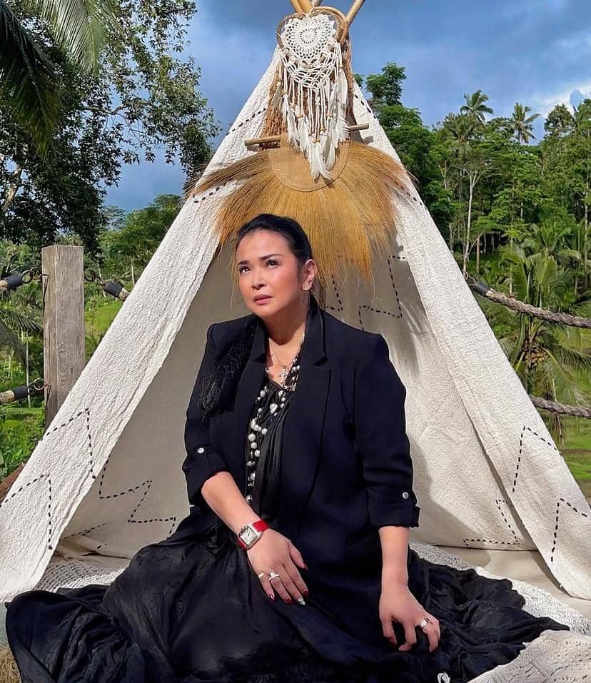 Seorang wanita berbusana hitam duduk di depan tenda putih berhias anyaman dan bulu, dengan latar pepohonan hijau dan langit biru.