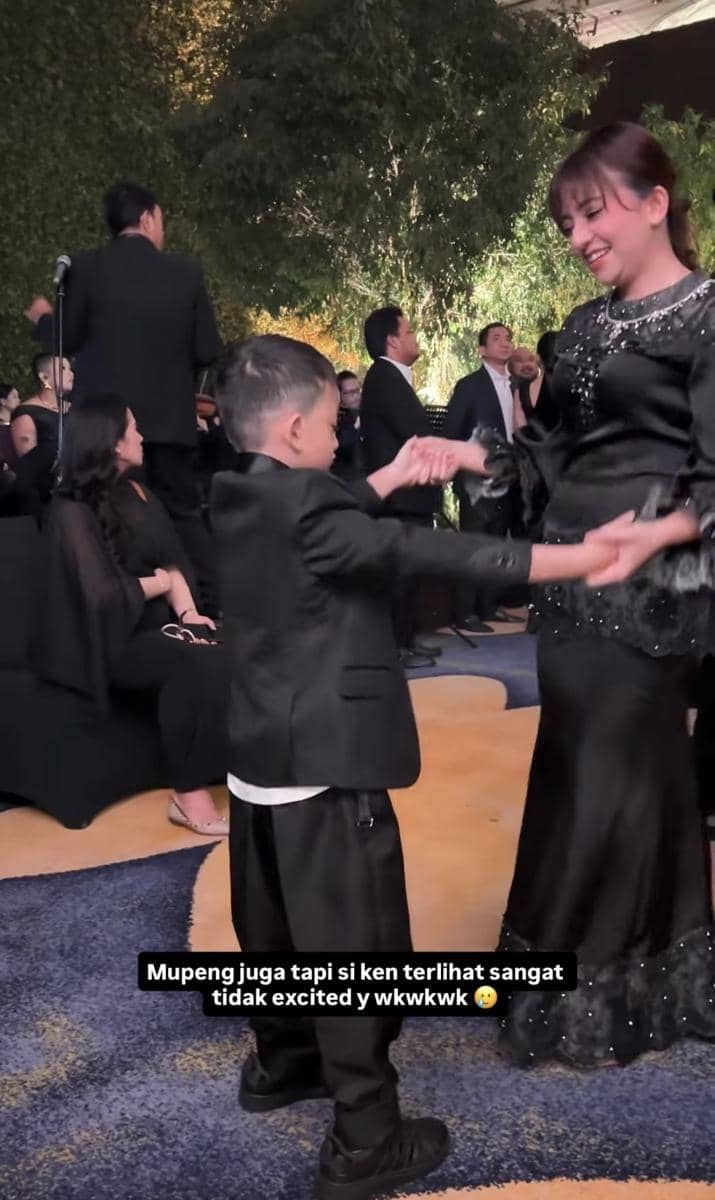 Seorang wanita mengenakan gaun hitam tersenyum sambil menari bersama anak laki-laki kecil berpakaian jas hitam di acara pernikahan.