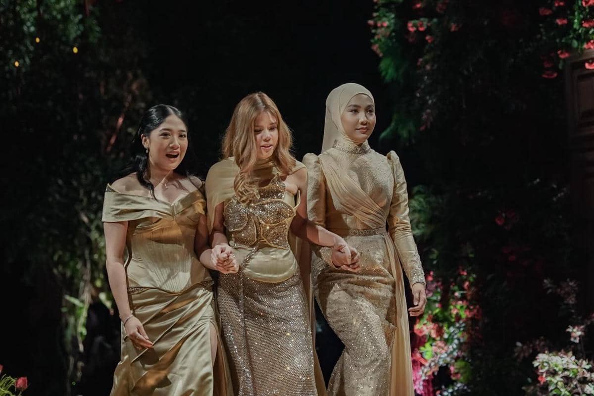 Tiga wanita mengenakan gaun berwarna emas berjalan bersama di acara resepsi dengan latar taman dan pencahayaan hangat.