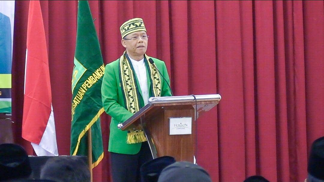 Seorang pria mengenakan pakaian hijau dan peci bermotif sedang berpidato di podium dengan latar bendera PPP dan tirai merah.