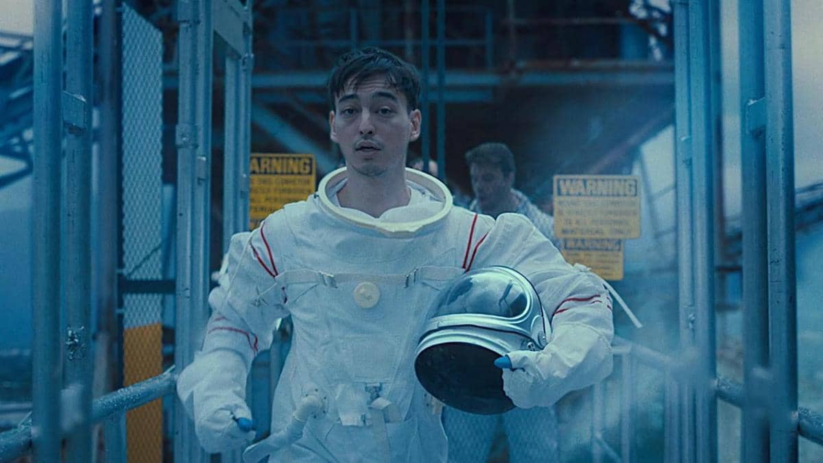 Seorang pria mengenakan pakaian astronot putih sambil memegang helm luar angkasa di area industri dengan suasana biru dingin.