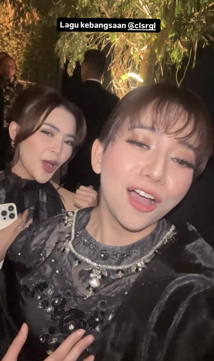 Dua wanita mengenakan gaun hitam berhias payet berpose sambil tersenyum di acara malam dengan pencahayaan hangat dan latar pepohonan.