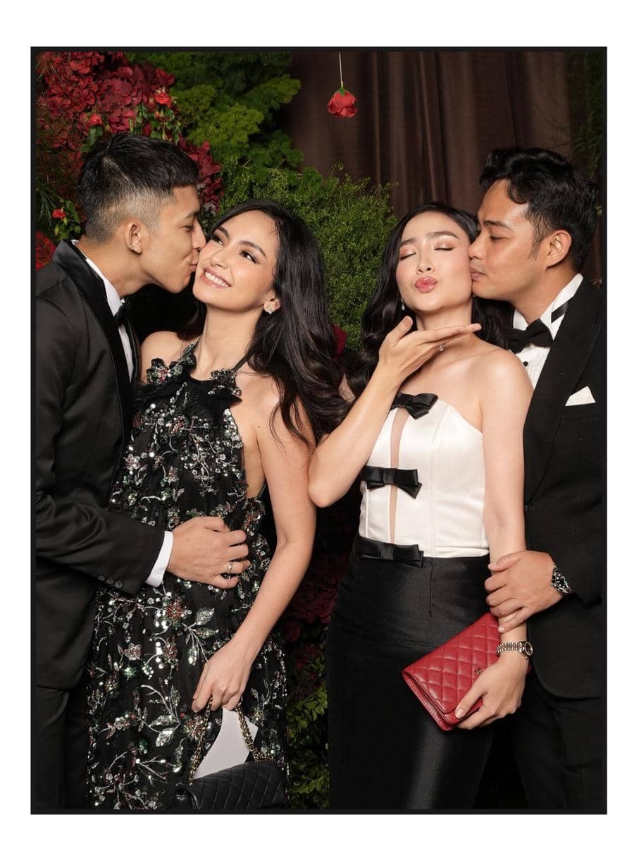 Dua pasangan berpakaian formal berpose mesra di acara resepsi pernikahan dengan latar dekorasi bunga dan tanaman hijau.