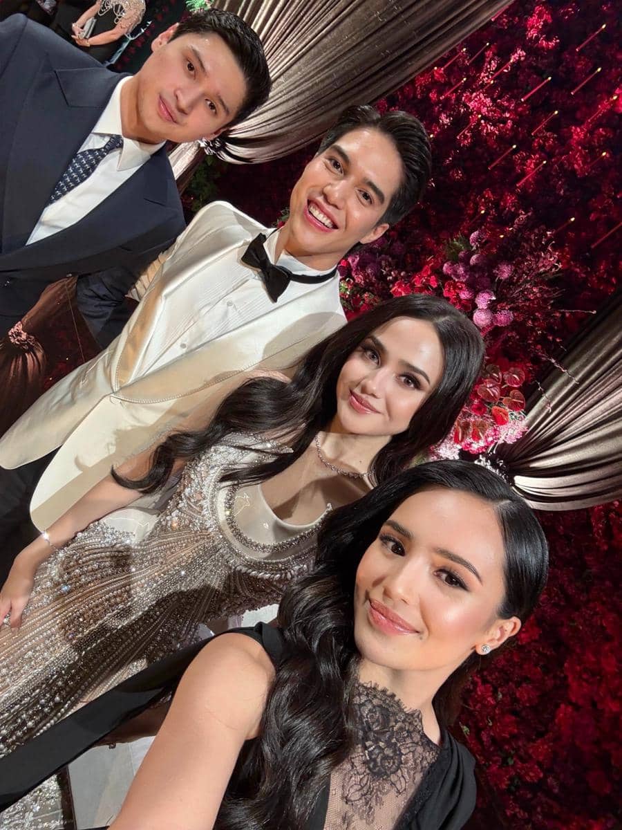 Empat tamu berpakaian formal berpose bersama di resepsi pernikahan dengan latar dekorasi bunga merah dan tirai elegan.