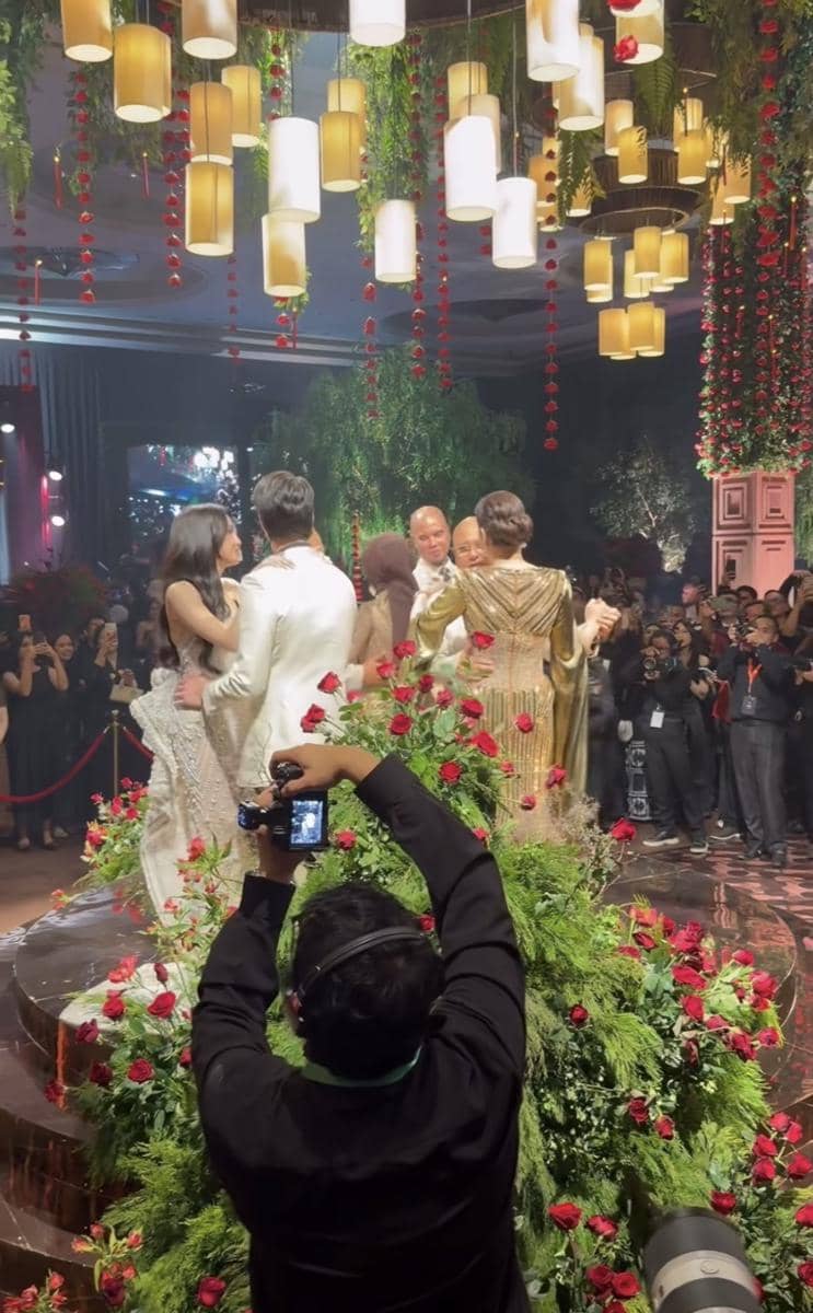 Pasangan mengenakan busana formal berdansa di panggung berhias bunga mawar merah dan lampu gantung dalam suasana pesta meriah.