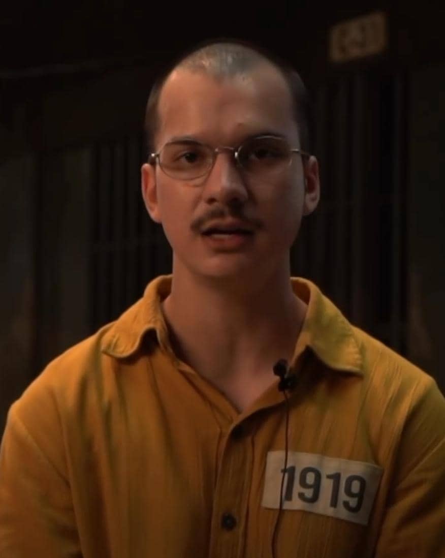 Seorang pria berkacamata mengenakan seragam tahanan berwarna oranye dengan nomor 1919 di dada, berada di ruangan redup.