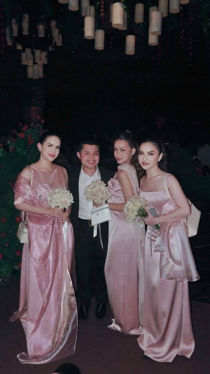 Tiga bridesmaid mengenakan gaun satin berwarna pink muda berpose bersama seorang pria di acara pernikahan dengan dekorasi bunga dan lampu gantung.