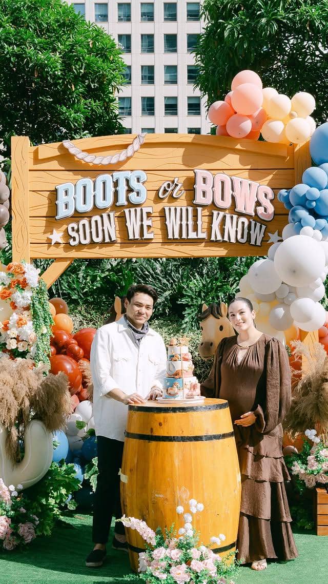 Pasangan berdiri di depan dekorasi pesta gender reveal bertema western dengan balon warna-warni dan tulisan 'Boots or Bows'.
