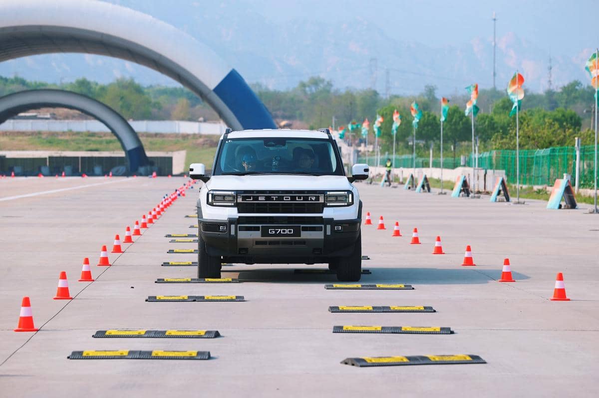 SUV Jetour G700 berwarna putih melaju di lintasan uji dengan deretan cone oranye dan rintangan kecepatan di area terbuka.