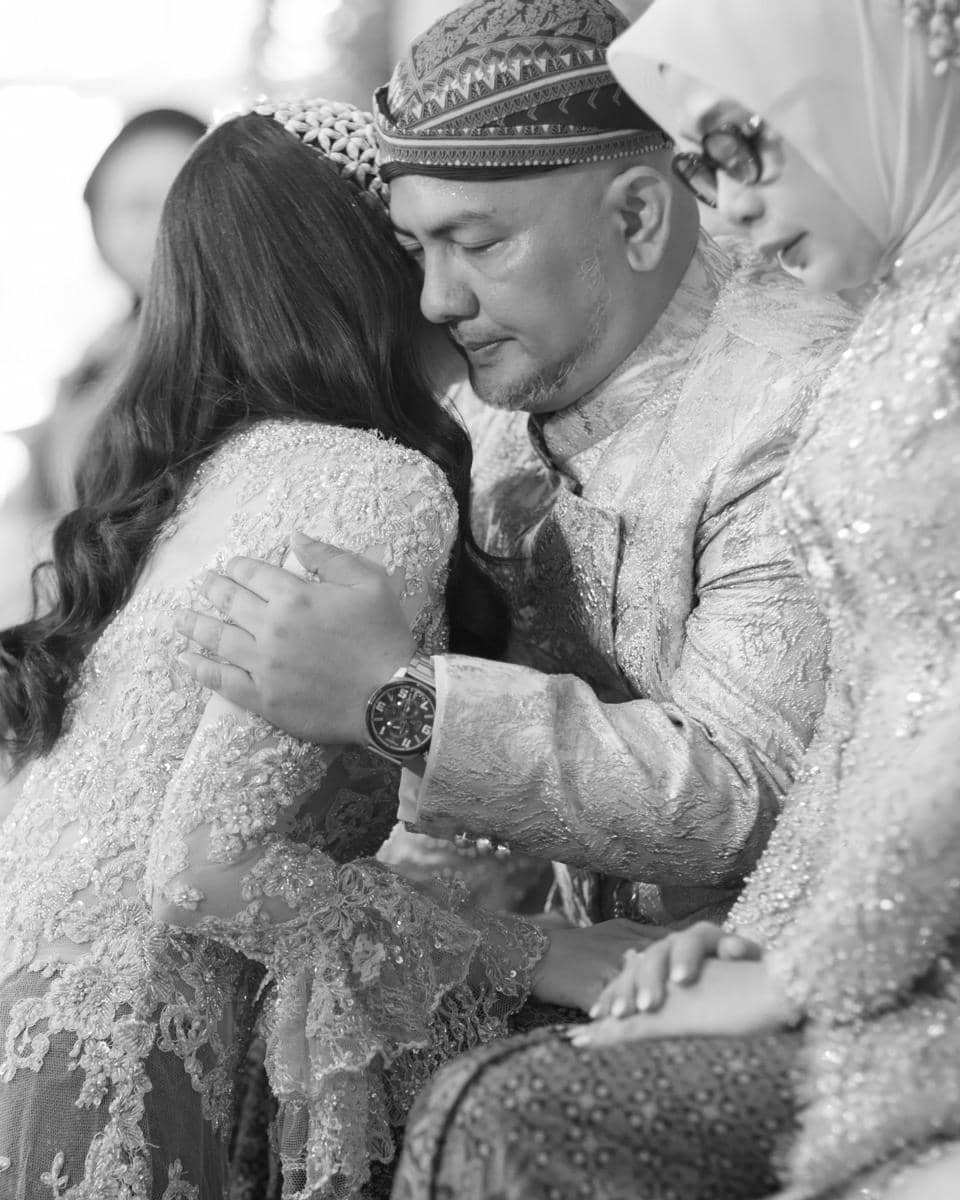 Seorang pria mengenakan pakaian adat berwarna senada memeluk seorang wanita berhijab dalam suasana haru di acara pernikahan.