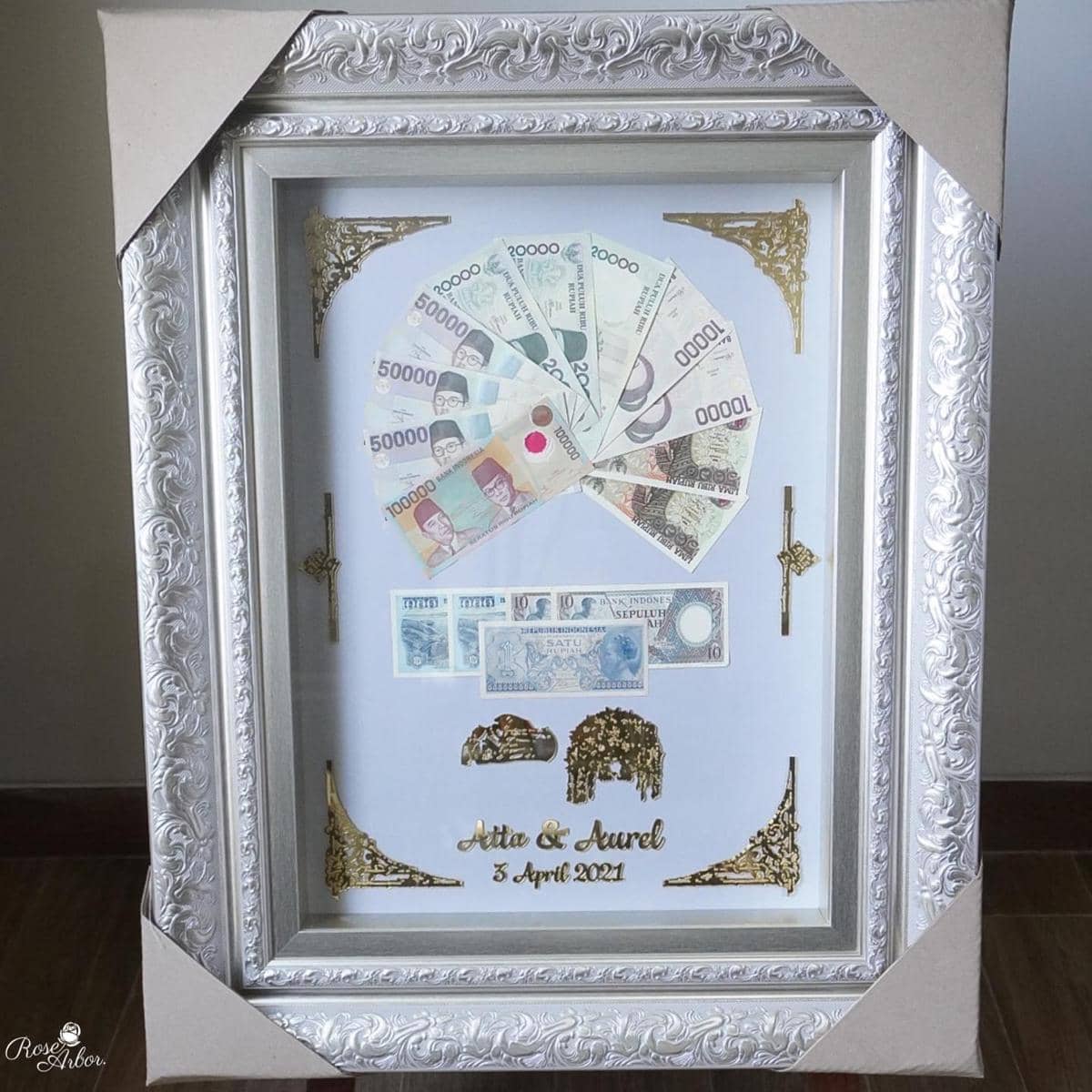 Bingkai mahar pernikahan berisi uang berbagai nominal dan mata uang asing dengan hiasan emas serta tulisan tanggal pernikahan.