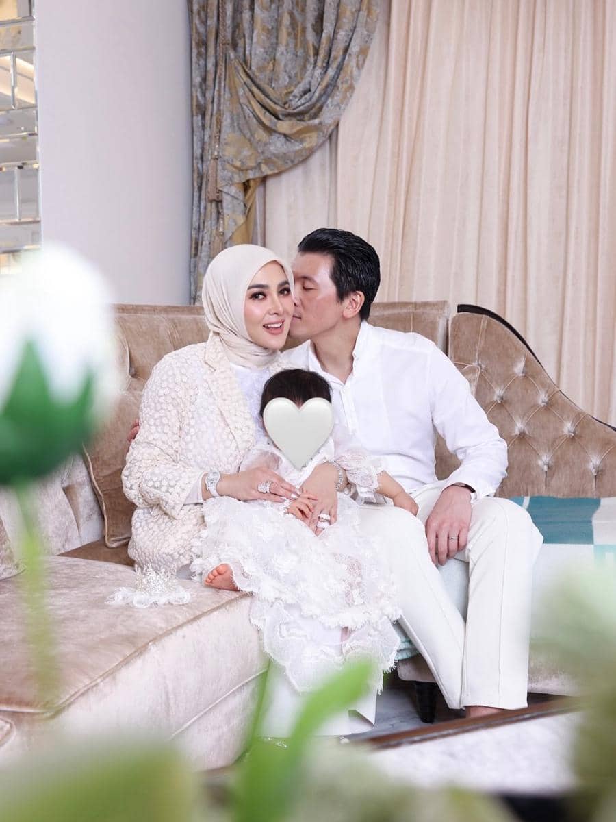 Syahrini dan Reino Barack berpakaian putih duduk di sofa bersama anak kecil yang wajahnya ditutupi bentuk hati di ruang tamu elegan.