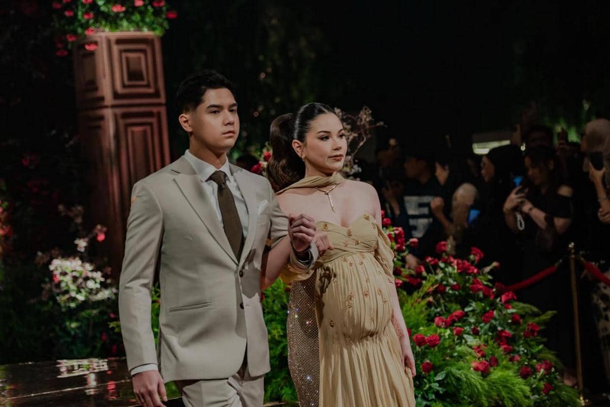 Sepasang tamu mengenakan busana formal berjalan di karpet resepsi dengan latar bunga merah dan dekorasi elegan di malam hari.