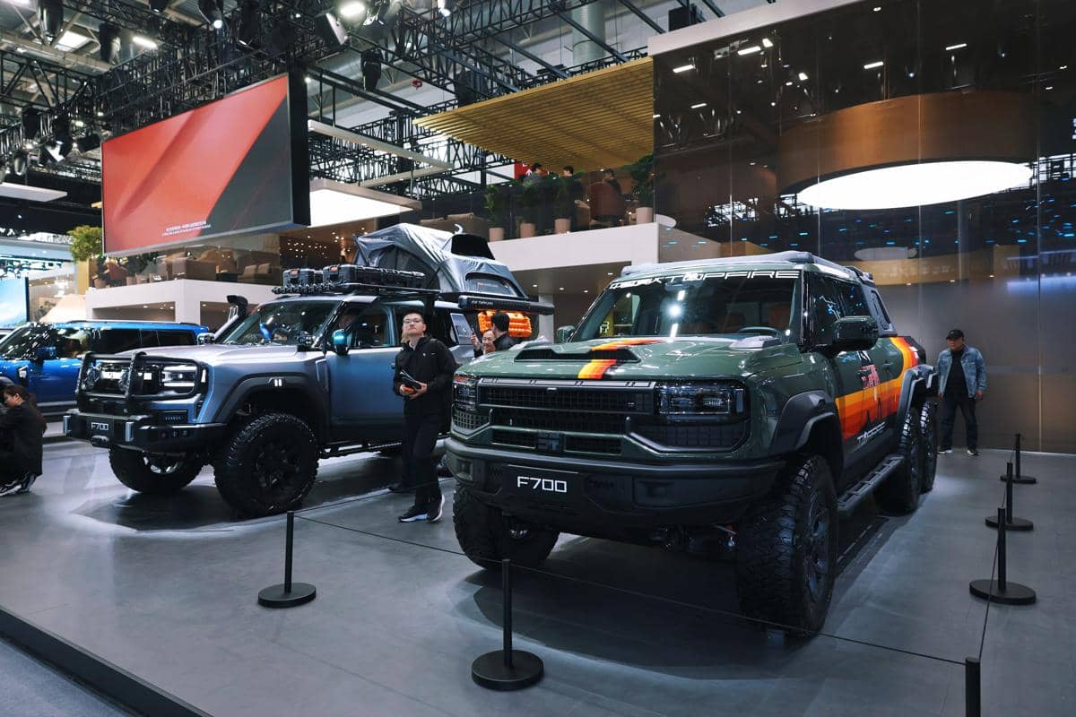 Dua unit Jetour G700 berwarna hijau dan biru dipamerkan di sebuah pameran otomotif dengan pencahayaan modern dan pengunjung di sekitarnya.