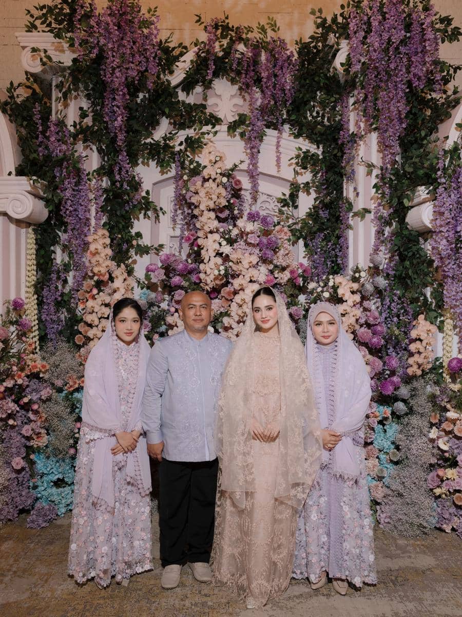 Seorang pria berdiri bersama tiga perempuan mengenakan busana tradisional berwarna pastel di depan dekorasi bunga ungu dan merah muda.