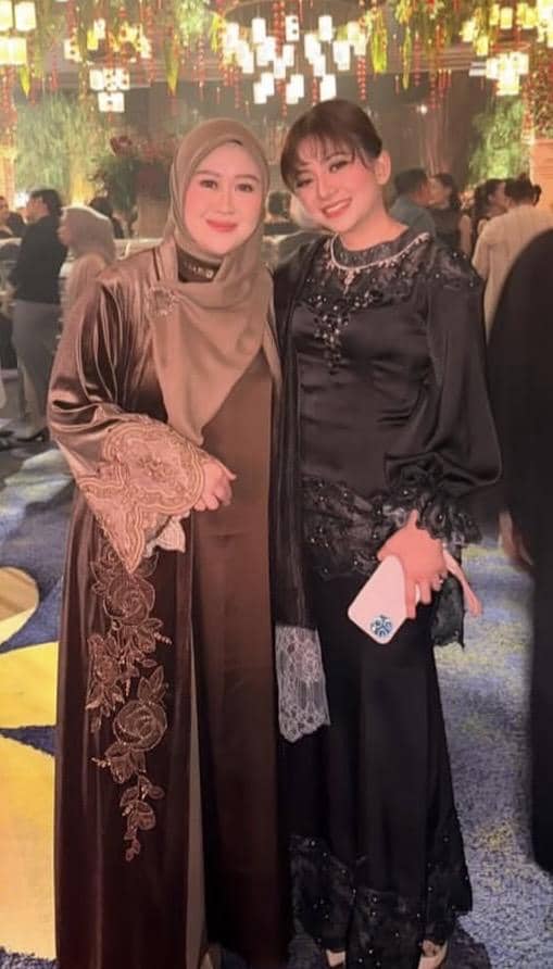 Dua wanita berpose bersama mengenakan busana pesta elegan berwarna cokelat dan hitam di acara pernikahan dengan dekorasi mewah.