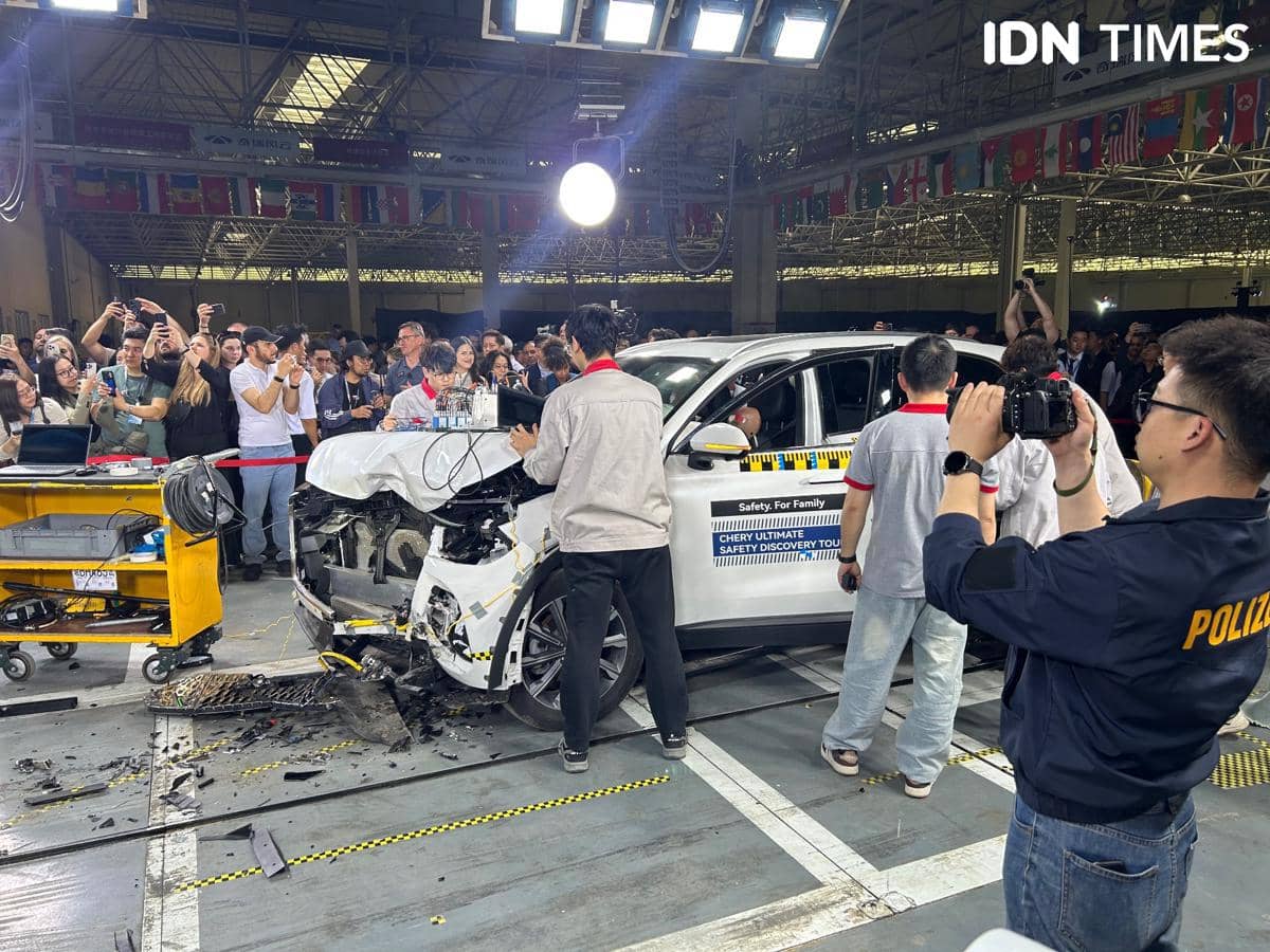 Mobil putih dengan bagian depan rusak setelah uji tabrakan dikelilingi teknisi dan penonton di area pengujian keselamatan.