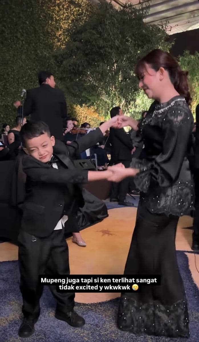 Seorang wanita bergaun hitam menari bersama anak laki-laki berpakaian jas hitam di acara pernikahan dengan latar taman malam hari.