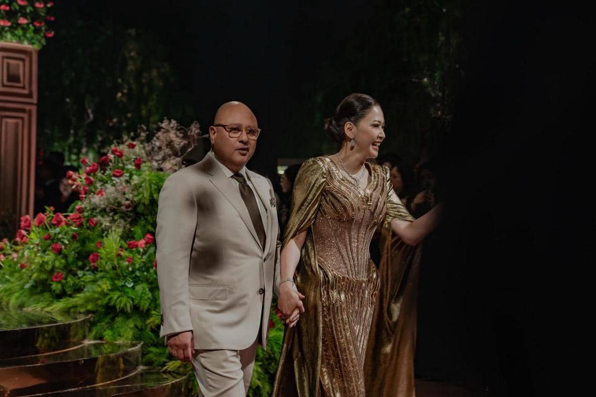 Sepasang tamu mengenakan busana formal elegan berwarna emas dan krem berjalan bergandengan tangan di resepsi pernikahan.