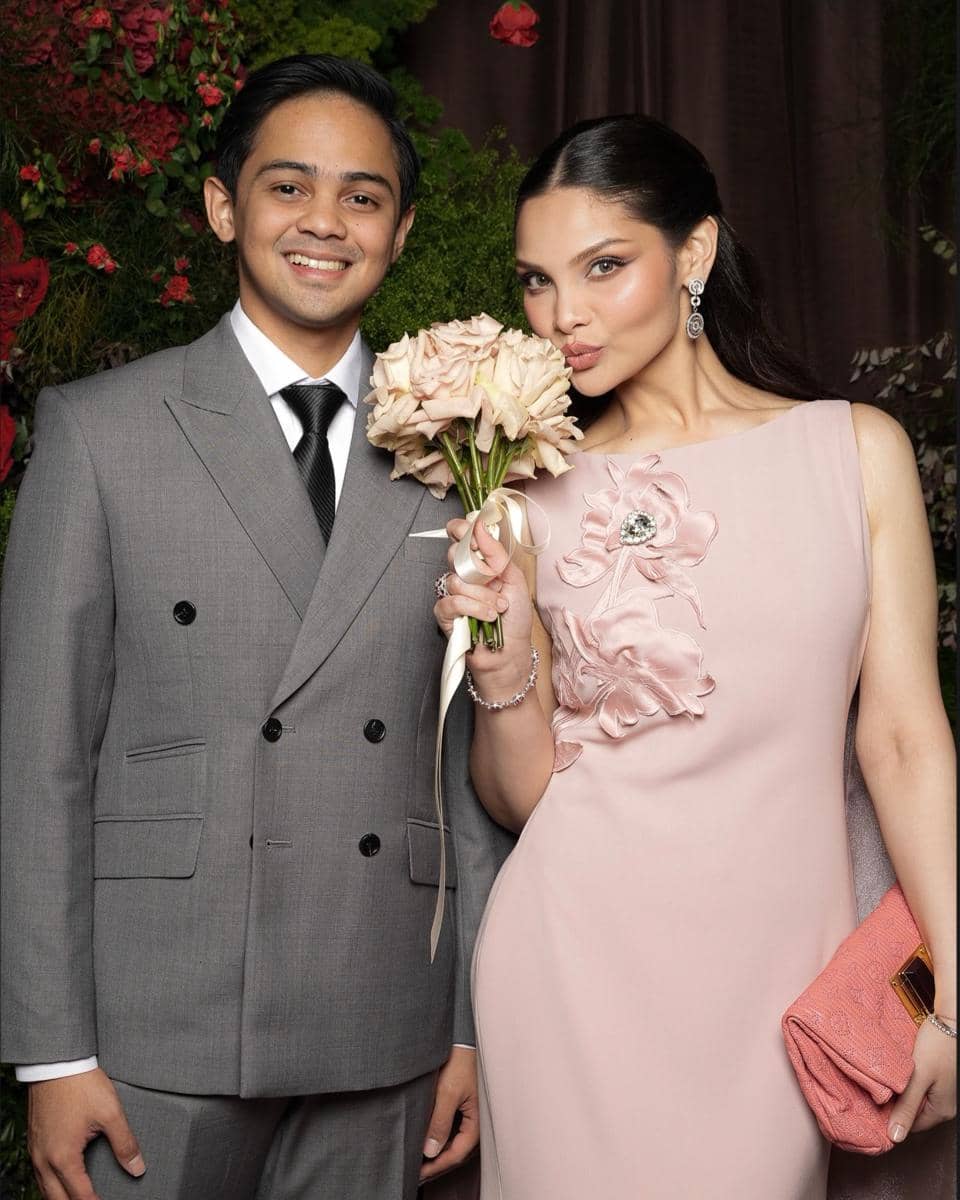 Seorang pria mengenakan setelan jas abu-abu berdiri di samping wanita bergaun merah muda yang memegang buket bunga mawar pucat.